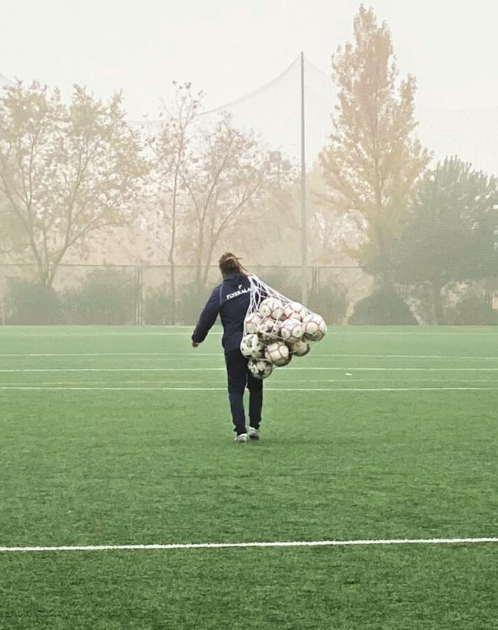 Entrenador de fútbol cargando una bolsa de balones, representando preparación, planificación y un entorno orientado al desarrollo.