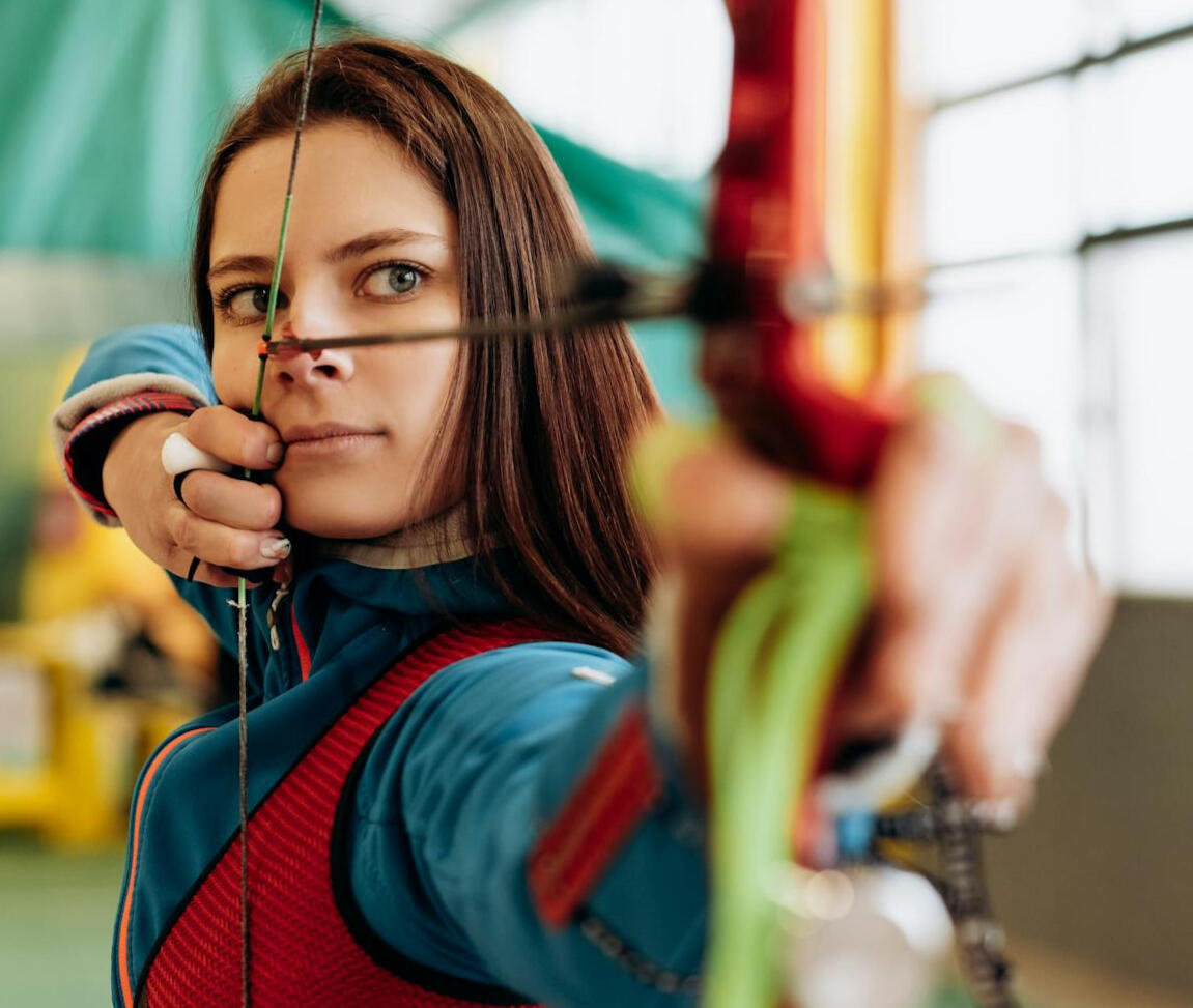 Arquera concentrada, con el arco en tensión y apuntando una flecha, representando concentración, control y entrenamiento mental de alto rendimiento.